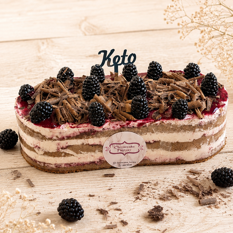 A keto chocolate cake with blackberries and chocolate shavings, decorated with a 'Keto' topper and a round logo sticker, set on a light wooden surface with scattered chocolate pieces and dried flowers.